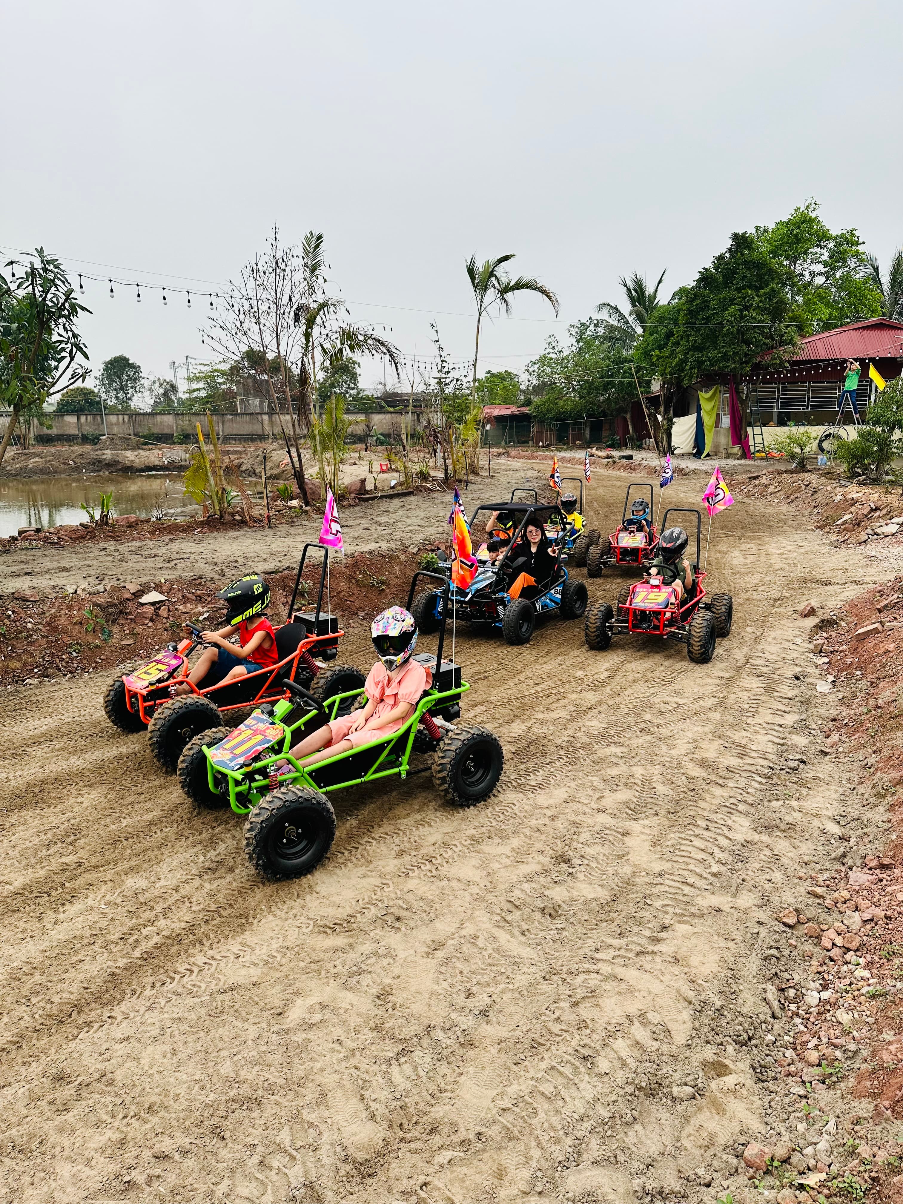 Đoàn xe địa hình mini xếp hàng trên đường đất, có cả trẻ em và người lớn đội mũ bảo hiểm